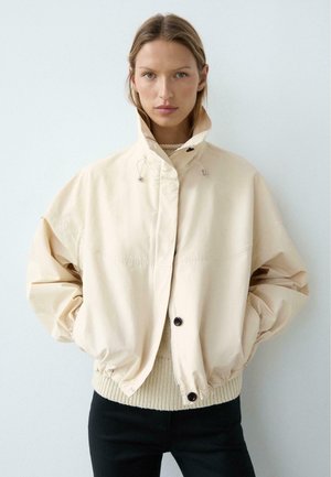 Femme portant une veste beige oversize à col haut, les mains dans les poches, sur un pull en maille crème et un pantalon noir, fond neutre.