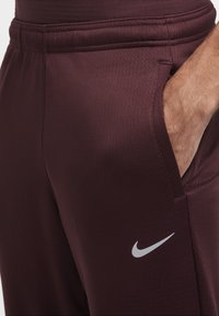 Bordowe spodnie sportowe z teksturowanej tkaniny, z bocznymi kieszeniami i szarym logo Nike na dolnej części nogawki. Elastyczny pas dla wygody.