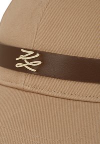 Tan cap made of textured fabric, featuring a brown leather band with a gold accent logo. Stitching visible along the brim.