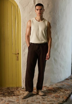 Ung mand iført en ærmeløs ribbet beige top, mørkebrune bukser med snøre og beige loafers stående på en stengulv ved en gul dør.