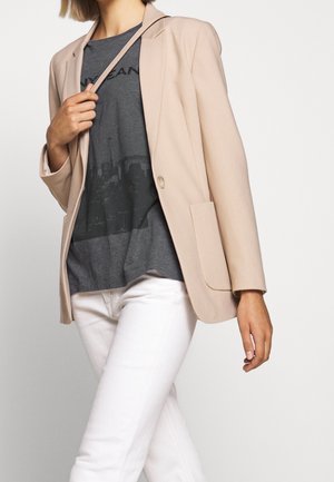 Kobieta ubrana w beżową marynarkę, ciemnoszarą koszulkę z grafiką i białe spodnie, trzymająca na ramieniu pasek beżowej torebki.