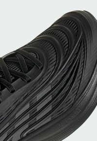 Zapato deportivo negro con parte superior texturizada con patrones ondulados elevados, cordones planos y suela de caucho resistente.