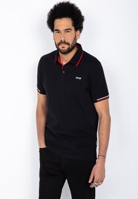 Sort polo skjorte med røde detaljer på kragen og ermene, med en tre-knapps placket og brodert logo på brystet. Laget av bomull.