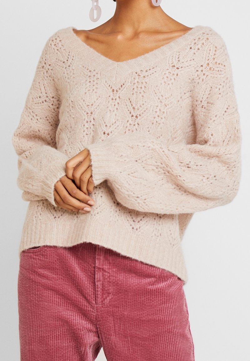 Pull en maille douce couleur crème avec motif en dentelle et col en V, porté avec un pantalon en velours côtelé rose taille haute et des boucles d'oreilles ovales pendantes.