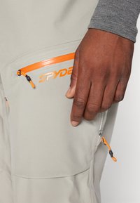 Ljusgrå byxor med orange dragkedjor och varumärkeslogotyp; har en ficka med dragband och texturerad tyg i närbild.