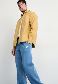 Dzeltena denima krekls ar pogām priekšā un kabatu; gaiši zili treniņbikses ar baltu Nike logo. Vienkārša fona, stāvoša poza.