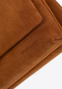 Brązowa torebka z zamszu o miękkiej fakturze, z delikatnym wytłoczonym logo. Projekt obejmuje proste linie i minimalistyczną estetykę.