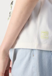 Hvit bomulls t-skjorte med rund hals og en subtil logo i neon gul på kanten, parret med lyse blå denimshorts.
