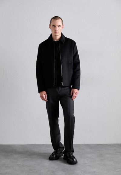 Black cropped jacket with a high collar, zip closure, and two side pockets, paired with dark gray slim-fit trousers and black shoes.