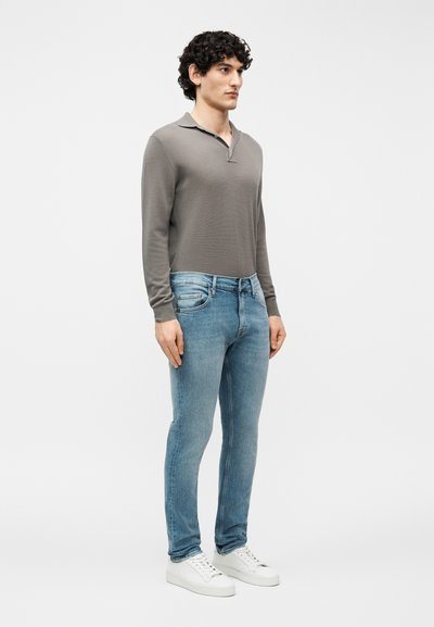 Νεαρός με σγουρά μαλλιά που φορά γκρι πόλο με μακριά μανίκια, ανοιχτό μπλε τζιν και λευκά αθλητικά παπούτσια, στέκεται μπροστά σε απλό φόντο.