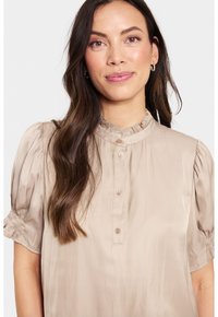 Blusa di seta beige con colletto alto arricciato e maniche corte a palloncino, con chiusura a bottoni e una texture liscia e lucida.