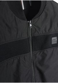 Crna bomber jakna s zatvaračem, rebrastim detaljima i glatkom teksturom, sa logotipom na prednjoj strani.