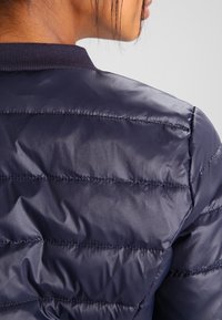 Marinblå puffjacka med kviltad design, slät textur och ribbad krage. Tyget verkar glänsande med jämnt fördelade sömnadsdetaljer.