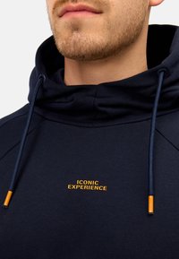 Mørkeblå hættetrøje med høj krave, der har et guld "ICONIC EXPERIENCE" logo og kontrastfarvede mørkeblå snore med orange spidser.