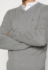Maglione grigio a scollo a V realizzato in tessuto morbido, con un piccolo logo sul lato sinistro del petto. Il design include maniche lunghe e polsini a costine.