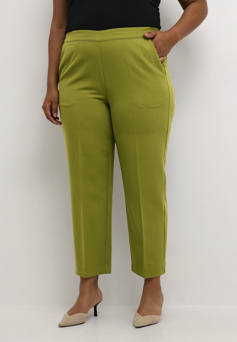 Personne portant un pantalon droit vert citron vert, un haut noir et des talons beiges à bouts pointus, debout avec une main dans la poche.