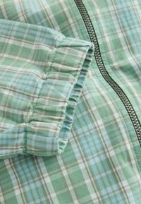 Tissu à carreaux vert et blanc avec détail de manchette élastique et fermeture éclair argentée visible sur un vêtement.