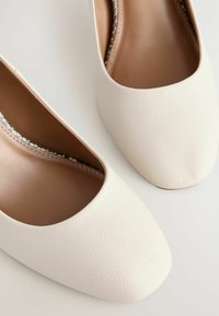 Zapatos cerrados de cuero blanco con un acabado texturizado y una plantilla suave beige. Presentan una ligera curva en la abertura para facilitar la entrada del pie.