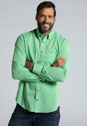 Grün karierter Button-down-Hemd mit Kragen, versehen mit braunen Knöpfen und einem kleinen Logo. Getragen zu dunklen Jeans. Heller grauer Hintergrund.