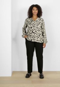 Schwarz-weißes Langarmbluse mit Blattmuster, kombiniert mit schwarzen Hosen und schwarzen Slippern. Die Bluse hat einen V-Ausschnitt und geraffte Ärmel.