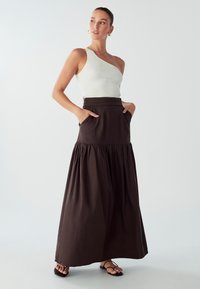 Top asimmetrico bianco a costine abbinato a una lunga e fluente gonna di cotone marrone con tasche e design a strati. Sandali neri completano l'outfit.
