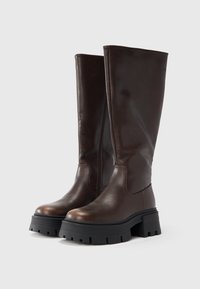 Rjavi usnjeni škornji do kolen z debelim črnim podplatom, sprednjo zadrgo in gladko teksturo, z minimalističnim dizajnom.