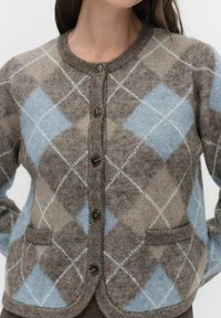 Kvinde iført en brun og blå argyle cardigan med knapper, to forlommer og ribbede kanter ved kraven og kanten.