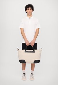 Geantă de voiaj bej din pânză, cu accente și curele din piele neagră; are un închidere cu fermoar și o formă rectangulară. Material texturat și mânere.