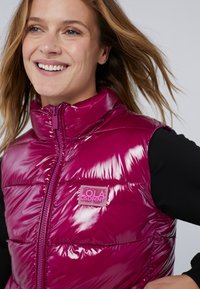 Fuchsia puffer vest met een hoge kraag, glanzende textuur, voorzakrits en een logopatch aan de linkerkant. Zwarte shirt daaronder.