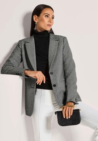 Grijze visgraat blazer met twee knopen, zwarte coltrui, witte broek en een kleine textuur zwart tas met een gouden handvat.