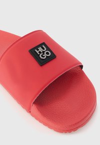 Rød rutsjebane sandal med en glat overflade, tekstureret fodseng og et sort rektangulært logo-patch med teksten "HUGO" på remmen.