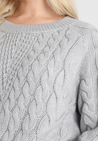 Pull gris en maille avec motifs torsadés, col rond et tissu texturé. Le design comprend des points en relief et torsadés.