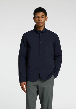 Giacca blu navy con colletto alto, bottoni a pressione frontali, maniche lunghe e una texture morbida, abbinata a pantaloni grigi.