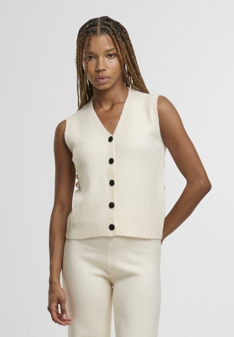 Cardigan senza maniche color crema con scollatura a V, caratterizzato da sei bottoni neri sul davanti e dettagli a coste sull'orlo. Materiale morbido e testurizzato.