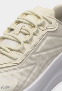 Κοντινό πλάνο μπεζ αθλητικού παπουτσιού με υφή υφάσματος, μαξιλαρωμένη γλώσσα, κορδόνια με θηλιά και λευκή σόλα, με το σήμα Calvin Klein στην πλευρά.