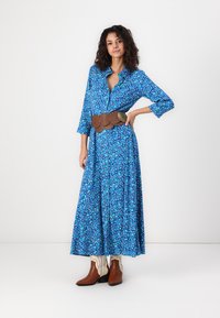 Μακρύ μανίκι, πουκάμισο φόρεμα σε μπλε με λουλουδάτα μοτίβα, τονισμένη μέση με καφέ ζώνη, συνδυασμένο με καφέ μποτάκια έως τον αστράγαλο και κρεμ παντελόνι.