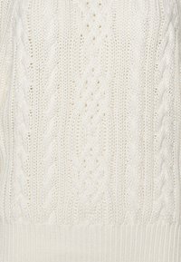 Stickad tröja i krämfärg med en texturerad kabelmönster, med ribbade kanter. Tyget verkar tjockt och mysigt, lämpligt för kallare väder.