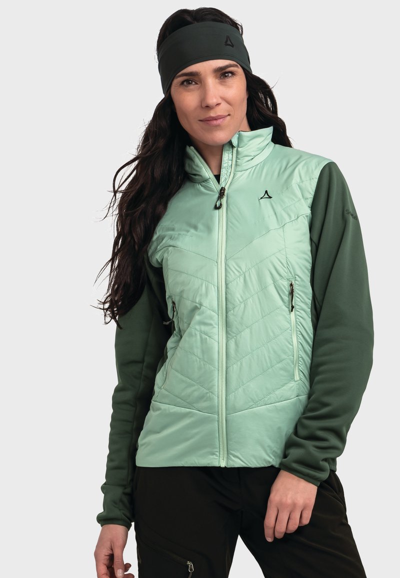 Hellgrüne gesteppte Jacke mit langen grünen Ärmeln, zwei Reißverschlusstaschen und hohem Kragen. Dazu wird ein passendes Stirnband getragen.