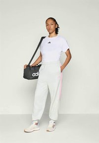 Baltā Adidas T-kreklā ar melno logo, pelēkās sporta biksēs ar rozā svītrām un baltos sporta apavos; tur tumsā Adidas soma.
