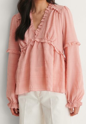 Frau trägt eine hellrosa Bluse mit langen Ärmeln, Rüschen und strukturiertem Stoff, kombiniert mit weißen Hosen, vor einem einfarbigen Hintergrund.