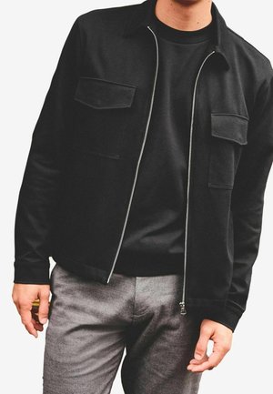 Homme portant une veste noire zippée avec des poches poitrine sur une chemise noire et un pantalon gris, debout les bras détendus le long du corps.