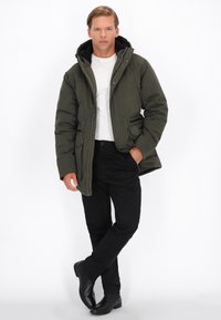 Jachetă puf de culoare verde cu glugă, detalii negre cu fermoar, purtată peste o cămașă albă și asociată cu pantaloni negri și cizme. Design simplu.