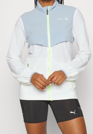 Persona con chaqueta con cremallera azul claro y blanca con cremallera neón, pantalones cortos negros de Puma, de pie con las manos entrelazadas al frente.