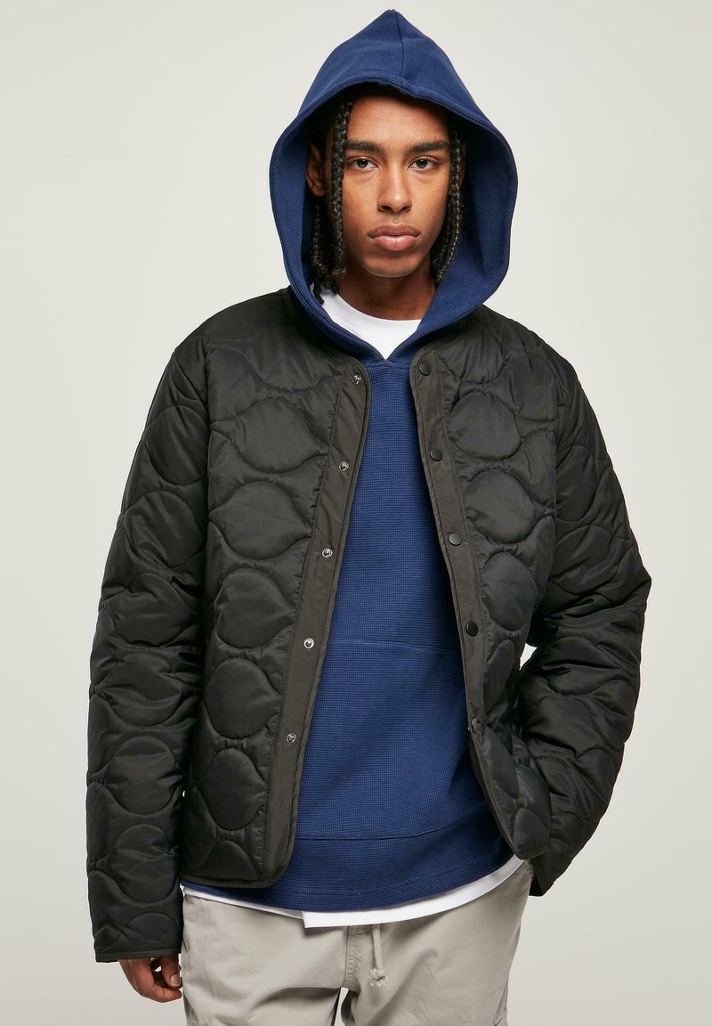 Svart kviltad jacka med glänsande yta och tryckknappar, bärs över en blå hoodie. Modellen har flätat hår.