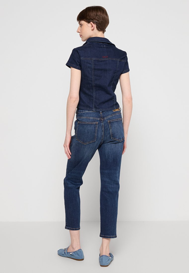 Giubbotto in denim scuro con design colletto, maniche corte e vita aderente. La parte posteriore presenta due tasche e un sottile dettaglio del logo.