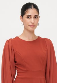 Rostfärgad klänning med en figursydd bodice, veckad långärm, rund halsringning och textilmaterial. Släta accentdetaljer i midjan.