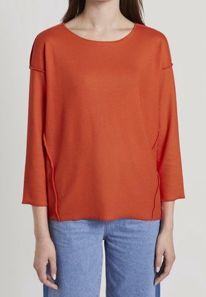 Femme portant un haut orange à manches longues ample avec un large décolleté et un jean bleu, montrée des épaules aux hanches.