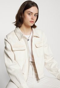 Jaqueta de ganga creme com dois bolsos no peito, fecho de correr à frente e colarinho pontiagudo. Usada por cima de uma blusa branca e acompanhada de calças de cores claras.