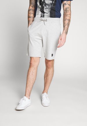 Shorts en coton gris clair avec un cordon de serrage à la taille, arborant un petit patch logo noir. Associés à des baskets blanches et une chemise à motifs.