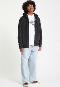 Sort hættejakke med lynlås lavet af blødt stof, med forlommer og snore, kombineret med løstsiddende lyseblå jeans og lilla sneakers.